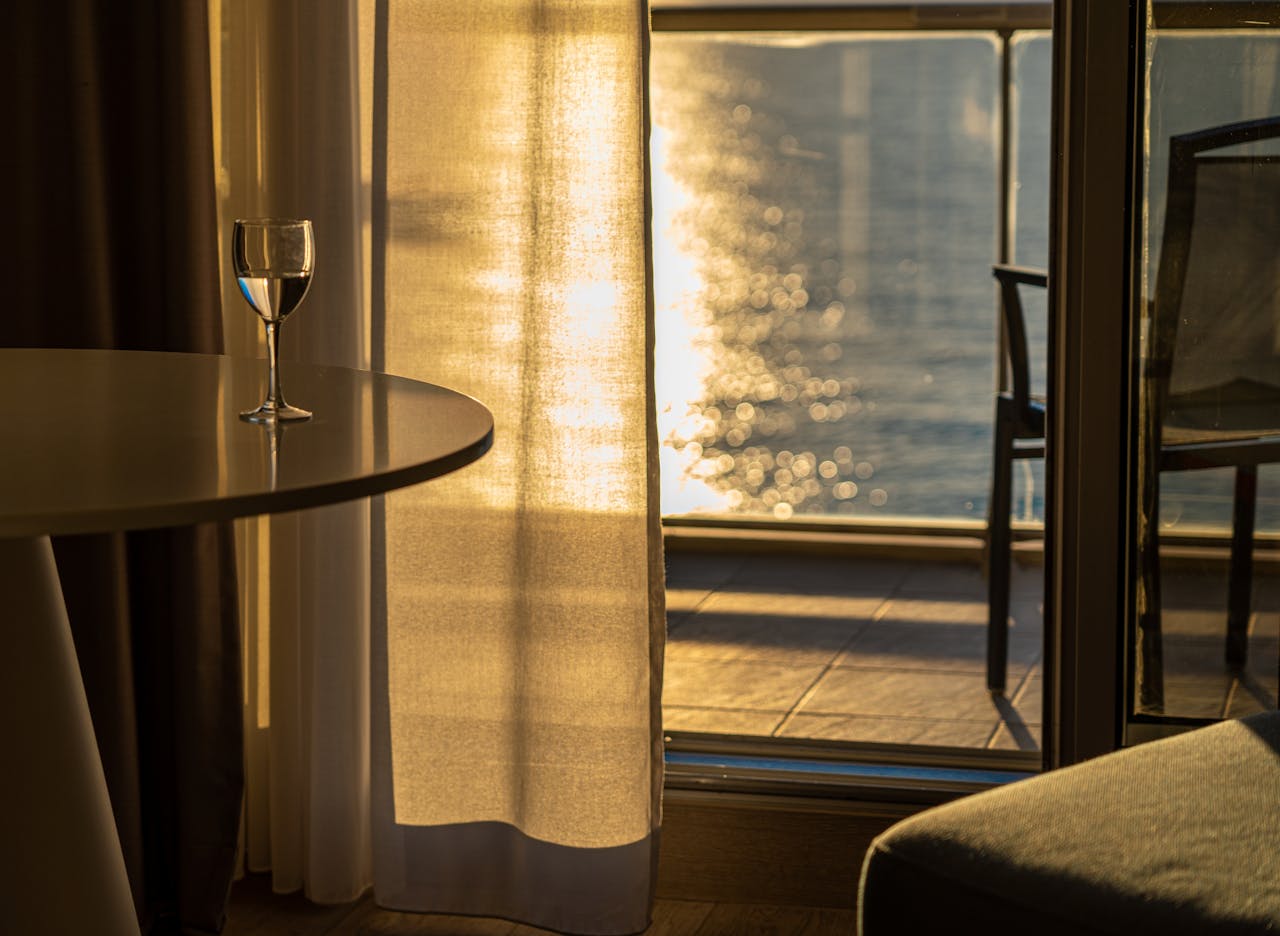 services-06 Sunlit room with a wine glass on table, overlooking a serene body of water from a balcony.
